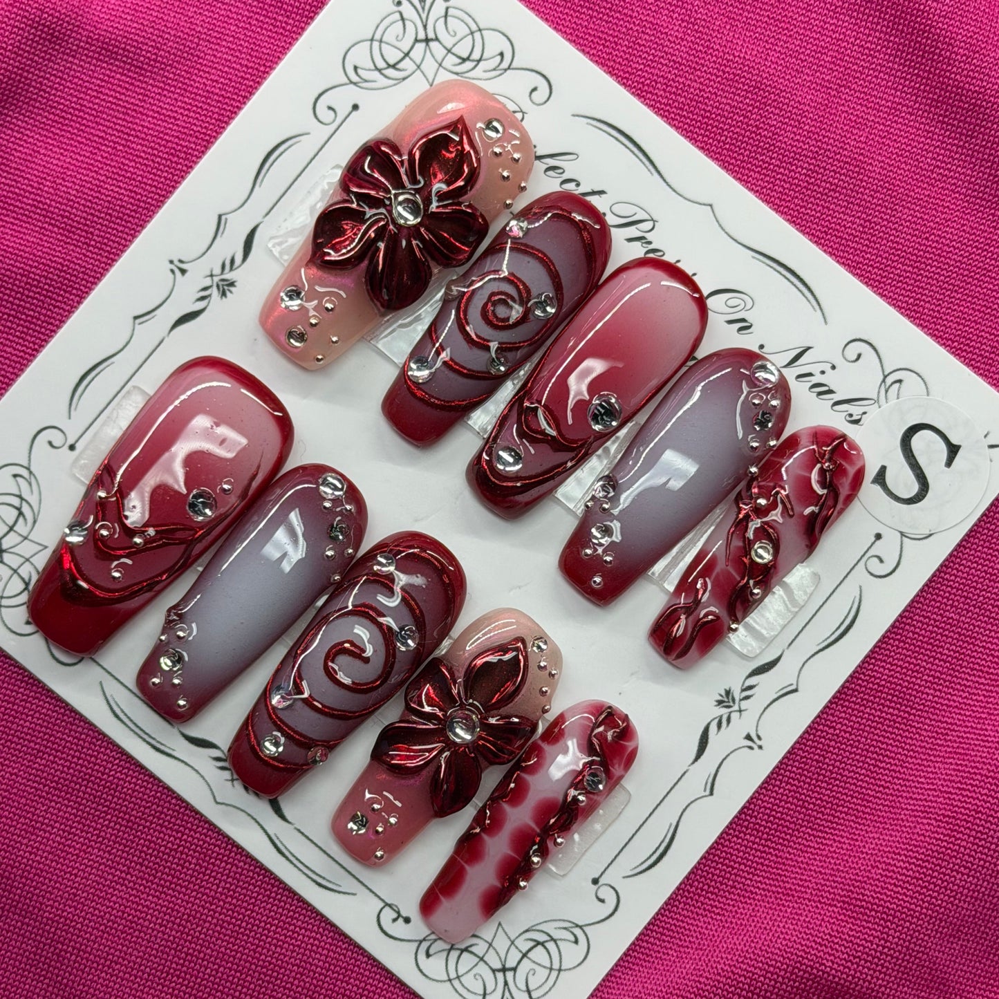 A set of 10 custom press-on nails displayed on a white card, featuring deep burgundy, metallic rose gold, and light lavender-gray colors with 3D floral designs and rhinestone accents, set against a textured magenta background.