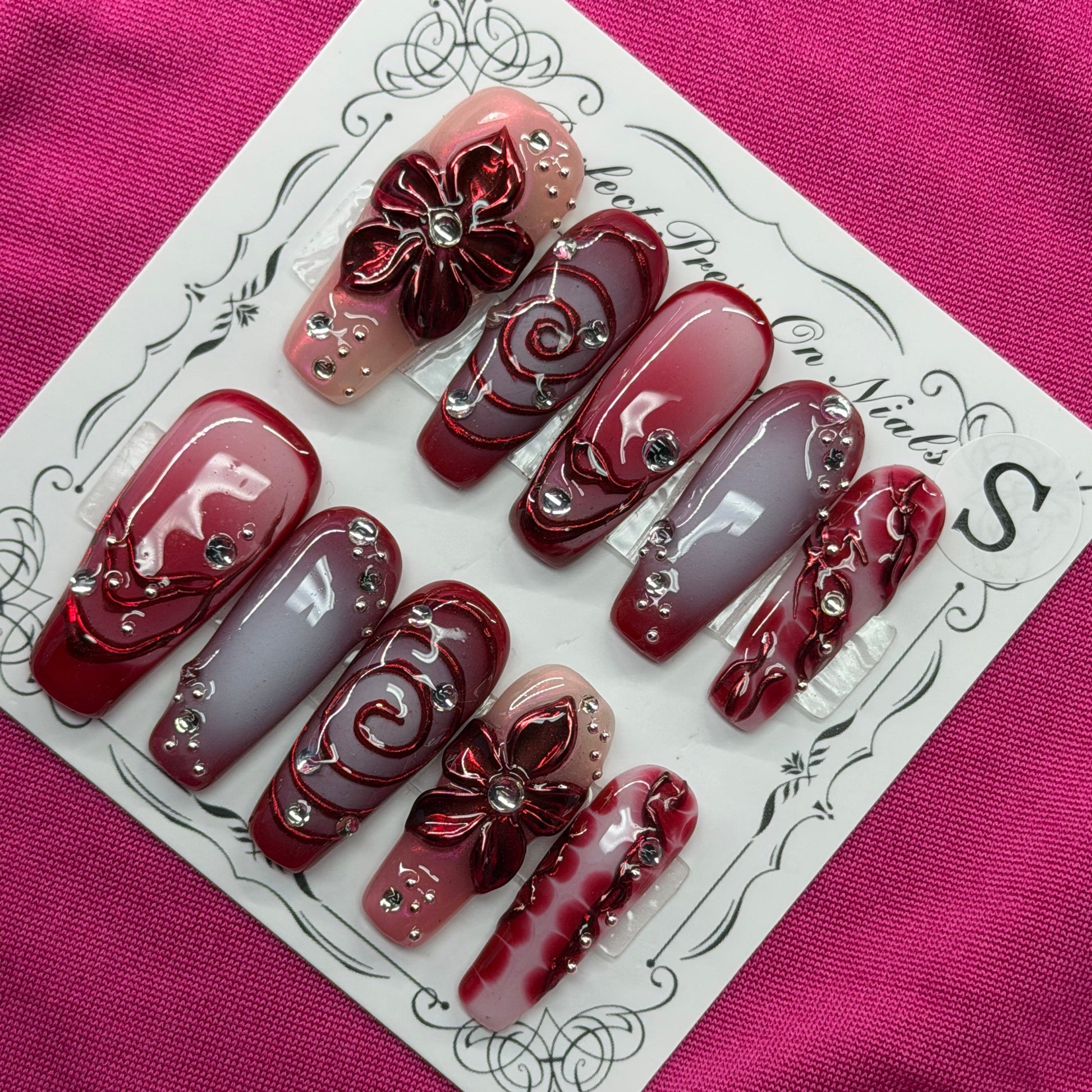 A set of 10 custom press-on nails displayed on a white card, featuring deep burgundy, metallic rose gold, and light lavender-gray colors with 3D floral designs and rhinestone accents, set against a textured magenta background.