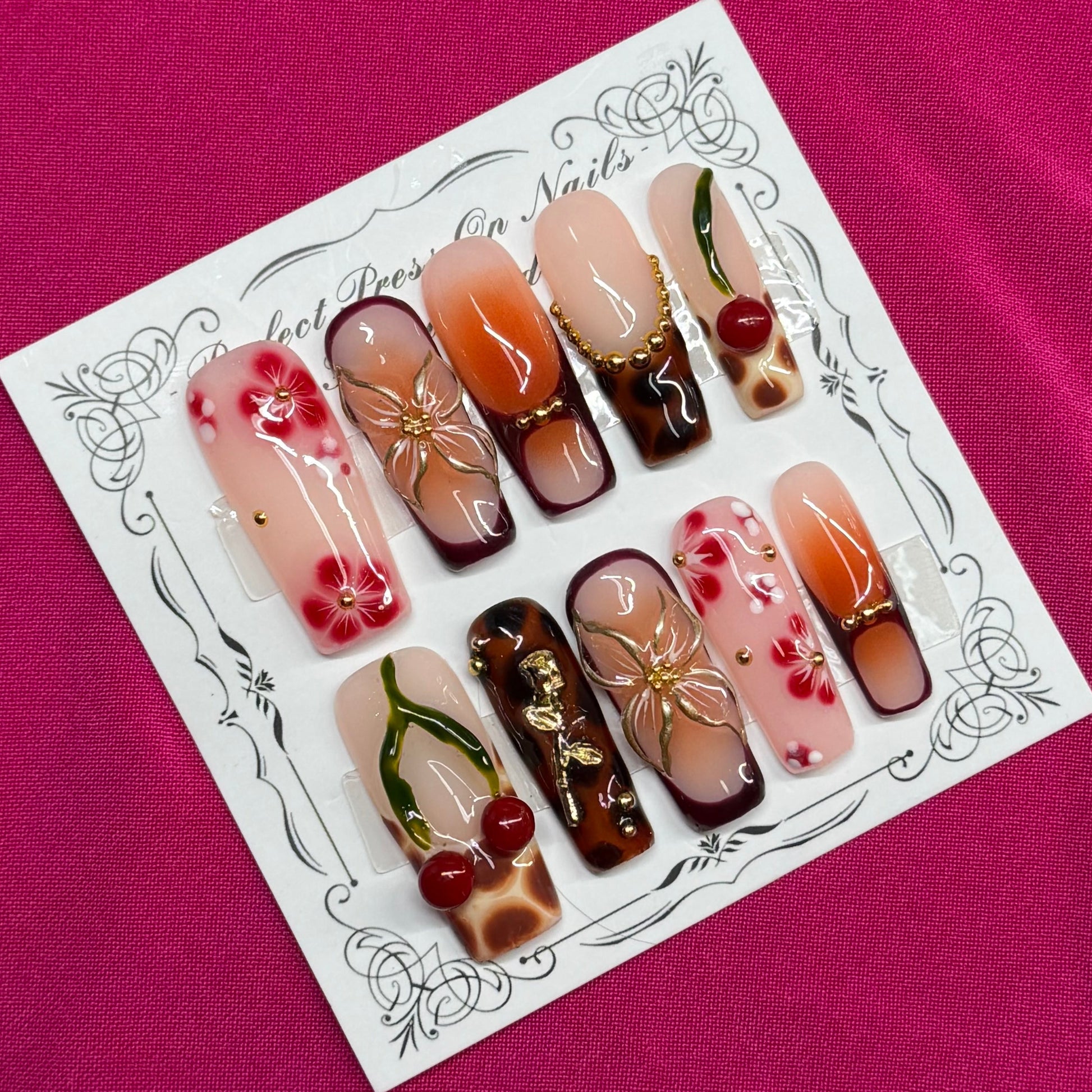 A set of decorative press-on nails displayed on a white card with an ornate border, placed on a bright pink fabric background. The nails feature various designs including floral patterns, cherry motifs, and gradient colors.
