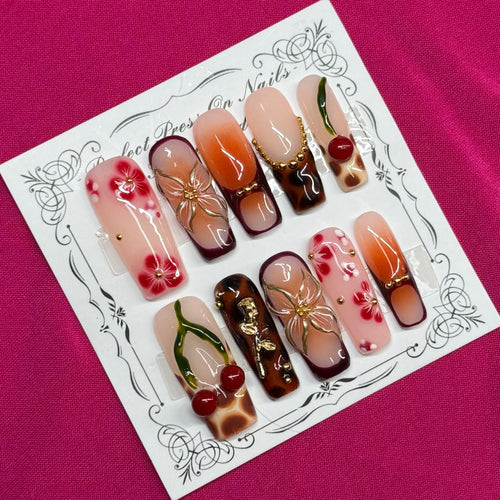 A set of decorative press-on nails displayed on a white card with an ornate border, placed on a bright pink fabric background. The nails feature various designs including floral patterns, cherry motifs, and gradient colors.