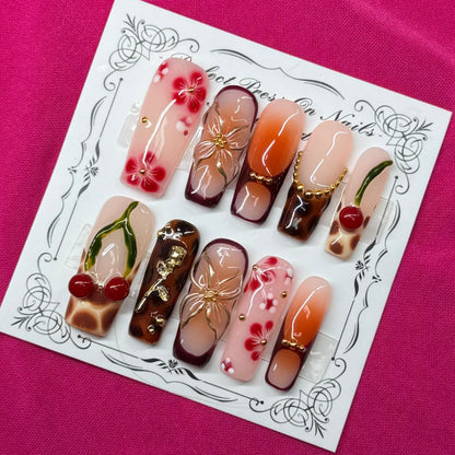 A set of 10 press-on nails displayed on a white decorative card, set against a vibrant magenta fabric background. The nails feature various designs including cherry motifs, gold leaf patterns, and dark brown glitter accents.