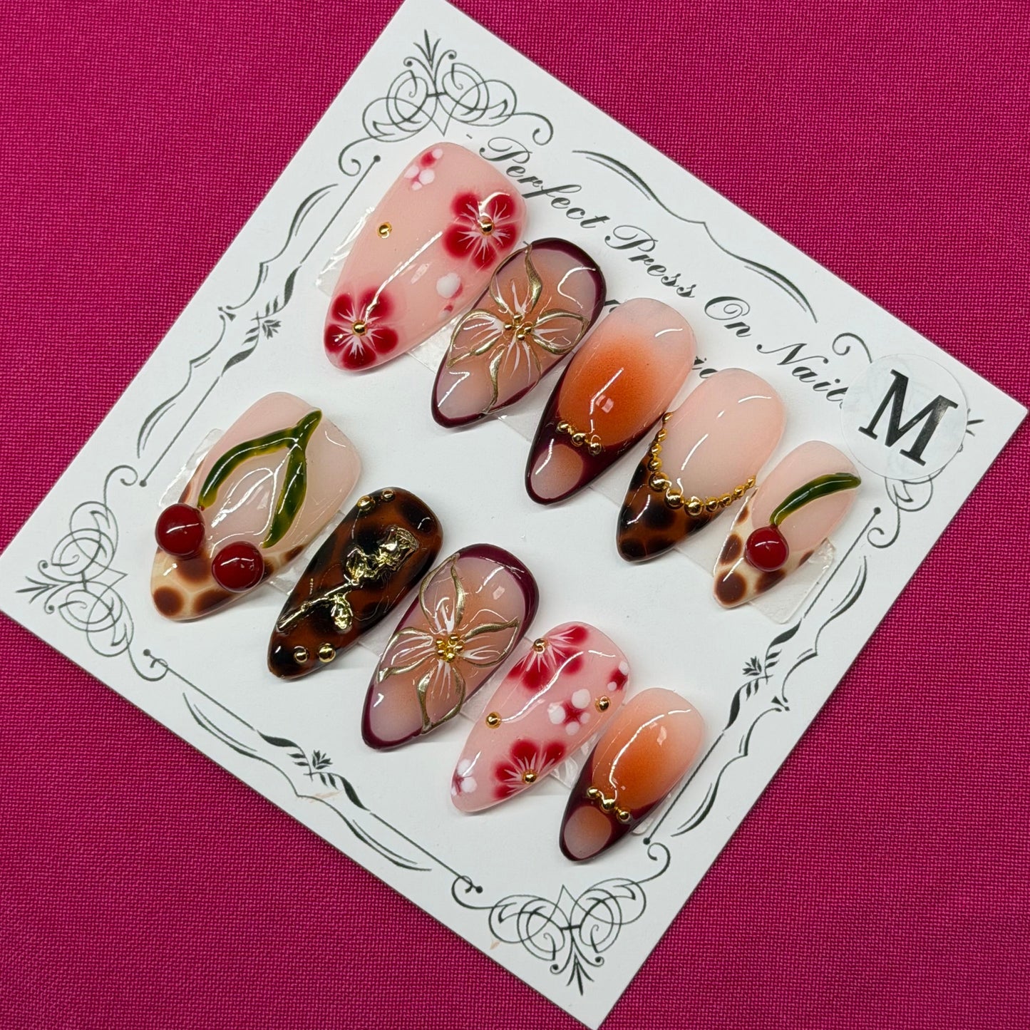 A set of decorative press-on nails displayed on a white card with ornate black detailing, placed on a solid magenta background. The nails feature various designs including floral patterns, cherry motifs, and metallic gold accents.