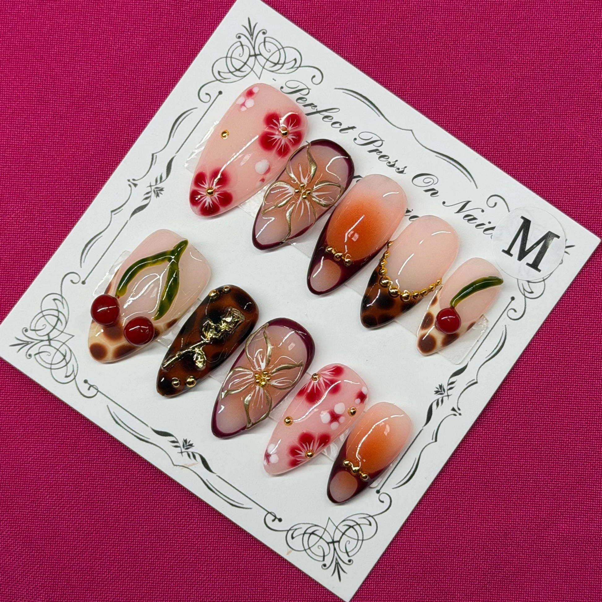 A set of decorative press-on nails displayed on a white card with ornate black detailing, placed on a solid magenta background. The nails feature various designs including floral patterns, cherry motifs, and metallic gold accents.