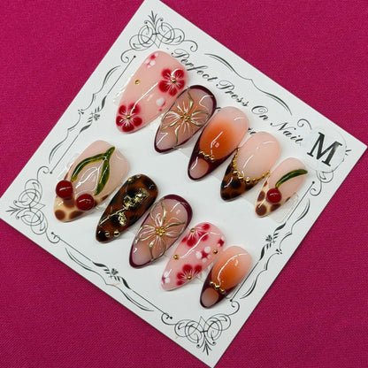 A set of decorative press-on nails displayed on a white card with ornate black detailing, placed on a solid magenta background. The nails feature various designs including floral patterns, cherry motifs, and metallic gold accents.