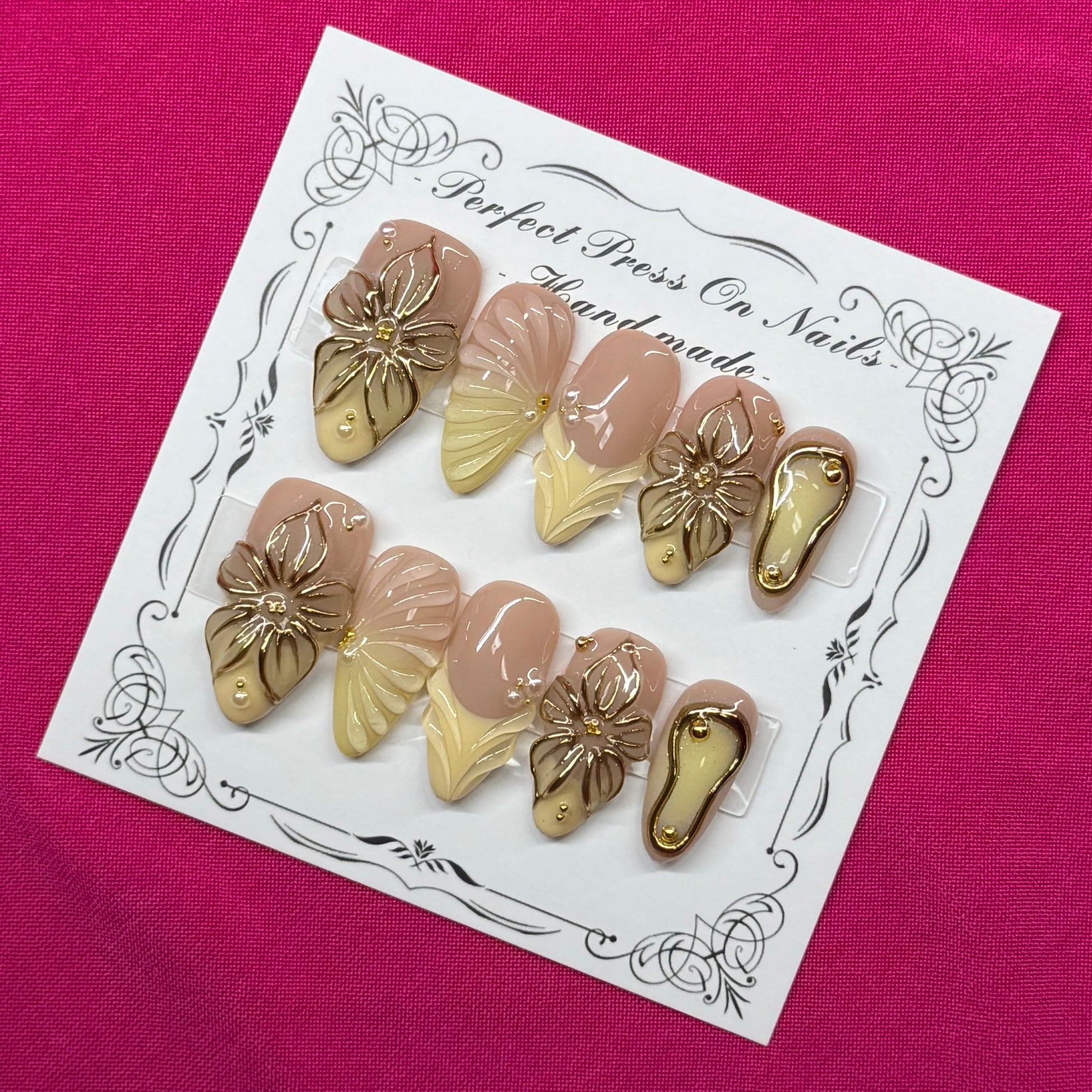 A set of 10 press-on nails displayed on a white decorative card, placed on a vibrant magenta fabric background. The nails feature a mix of metallic gold, champagne beige, and soft pink tones.