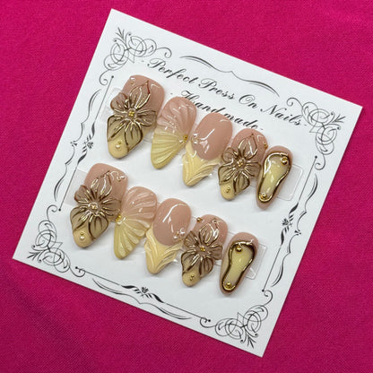 A set of 10 press-on nails displayed on a white decorative card, placed on a vibrant magenta fabric background. The nails feature a mix of metallic gold, champagne beige, and soft pink tones.