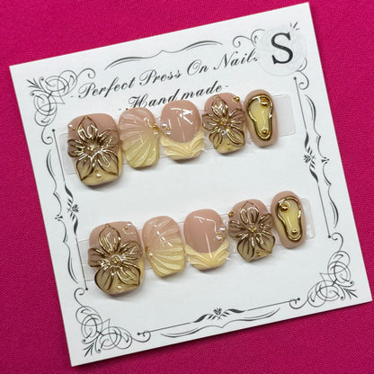 A set of 10 press-on nails displayed on a white card, featuring a mix of metallic gold, champagne, and soft pink tones, laid flat on a vibrant magenta fabric background.