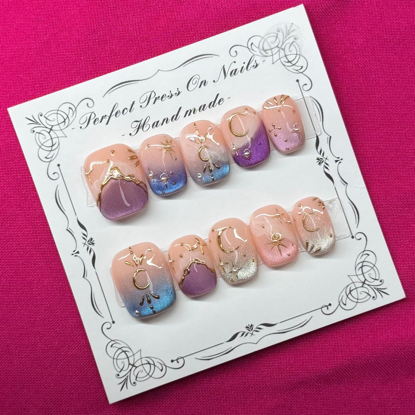 A set of handmade press-on nails displayed on a white card, placed on a vibrant magenta fabric background. The nails feature pastel pink, purple, and blue gradient designs with gold glitter accents.