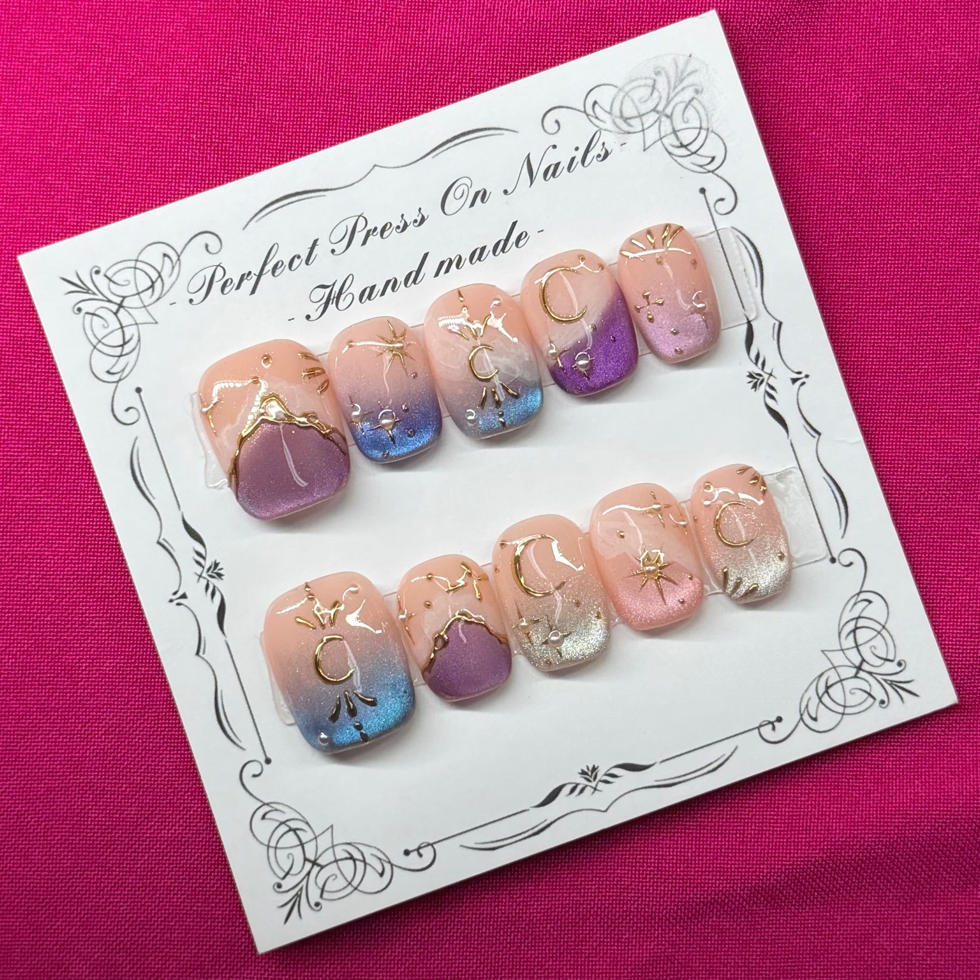 A set of handmade press-on nails displayed on a white card, placed on a vibrant magenta fabric background. The nails feature pastel pink, purple, and blue gradient designs with gold glitter accents.