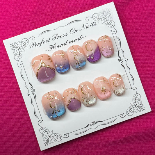 A set of handmade press-on nails displayed on a white card, placed on a vibrant magenta fabric background. The nails feature pastel pink, purple, and blue gradient designs with gold glitter accents.