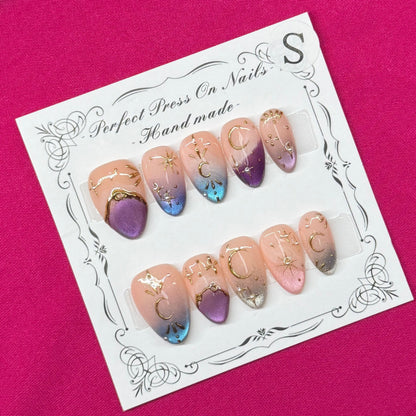 A set of decorative press-on nails displayed on a white card, placed on a bright pink fabric background. The nails feature a peach-to-purple gradient with blue glitter accents and gold foil details.