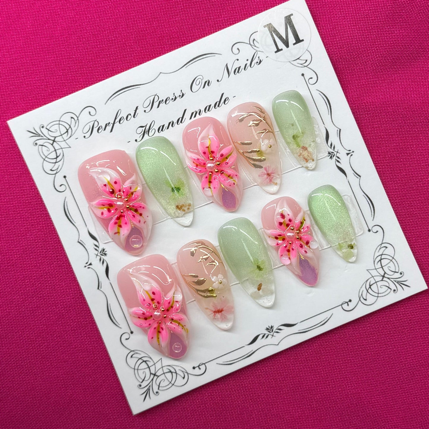 A 10-piece set of press-on nails on a white display card, featuring pastel green, pink, and gold glitter designs with floral details, placed on a vibrant magenta fabric background.