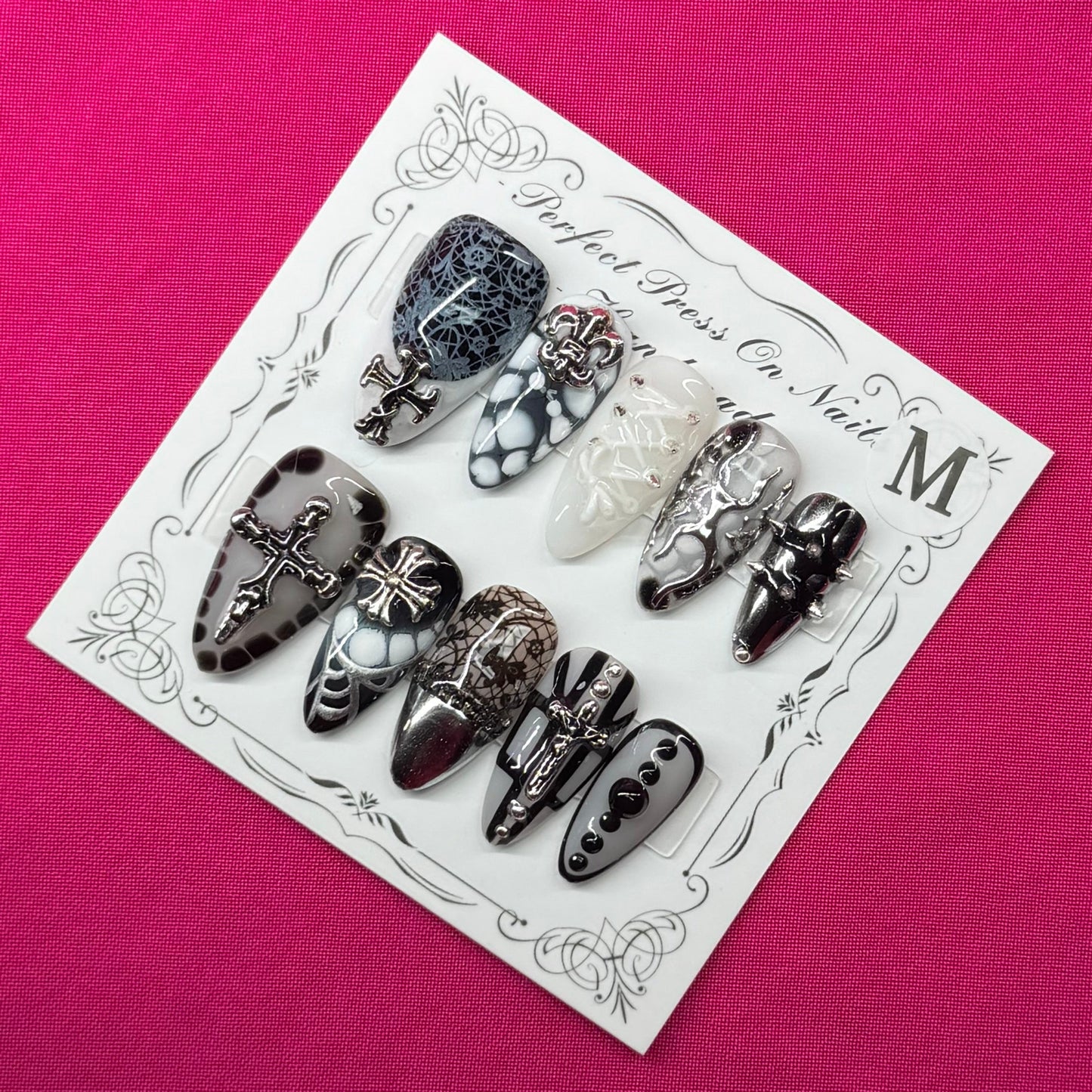 A set of decorative press-on nails on a small white card, placed on a bright pink fabric background. The nails feature black, white, and gray patterns including lace, glitter, and floral designs.
