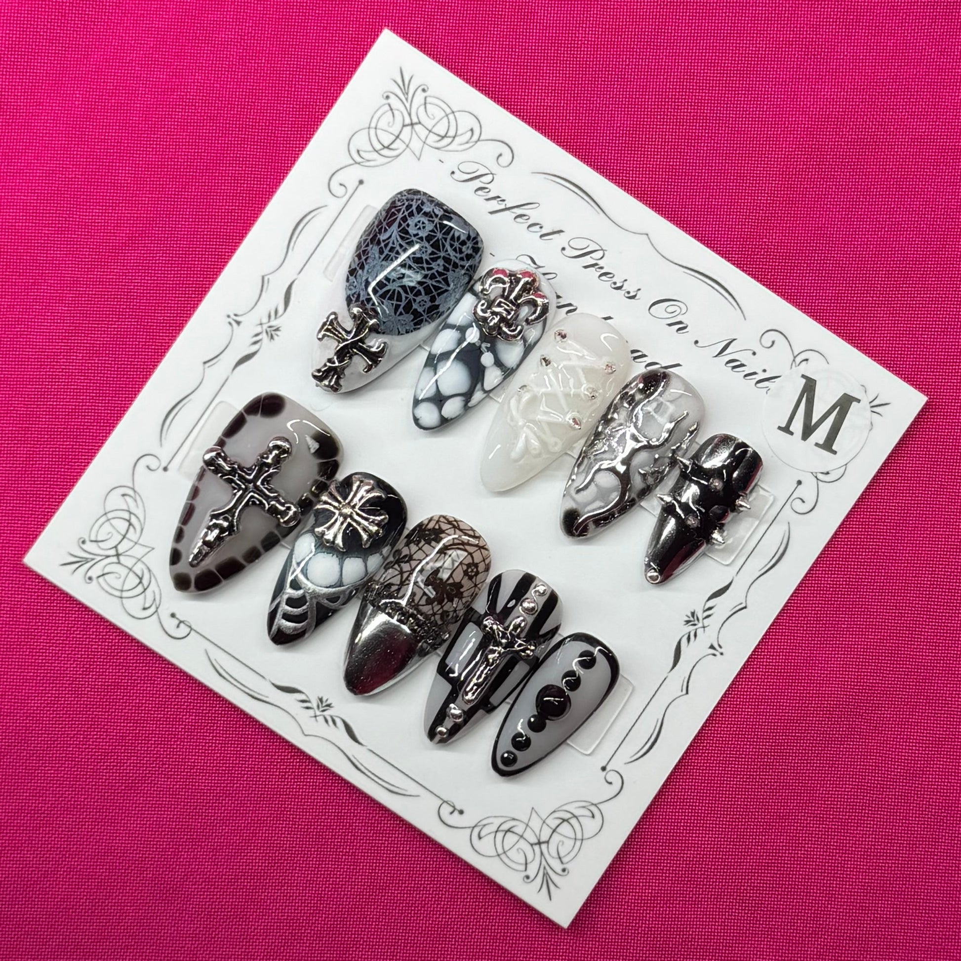 A set of decorative press-on nails on a small white card, placed on a bright pink fabric background. The nails feature black, white, and gray patterns including lace, glitter, and floral designs.