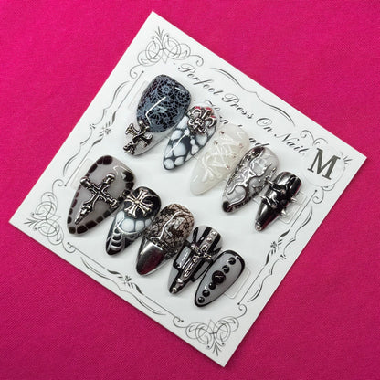 A set of decorative press-on nails on a small white card, placed on a bright pink fabric background. The nails feature black, white, and gray patterns including lace, glitter, and floral designs.